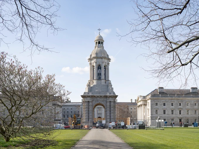 Alt Text: View of Trinity College Dublin featuring the front facade with the iconic campanile under a clear sky, surrounded by trees and greenery. Some construction equipment is visible in the foreground.