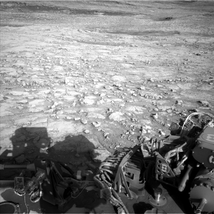 A black-and-white view from the Curiosity rover shows a rocky, textured Martian surface with scattered slabs and ridges. The rover’s mechanical components and wiring are visible in the foreground, along with its shadow stretching across the lower left of the frame. The horizon rises in the distance with layered terrain.