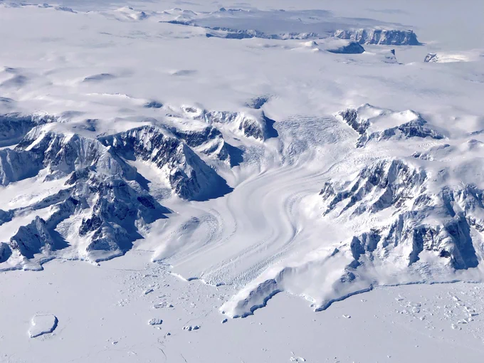 An aerial photograph of a glacier in mountainous terrain, flowing intoa frozen sea. There are bare rock peaks to the left of the image, and a few to the right, and a glacier with winding ridges cuts through them. At the bottom of the image is a flat white surface where ice covers the sea, broken up by icebergs trapped in it.