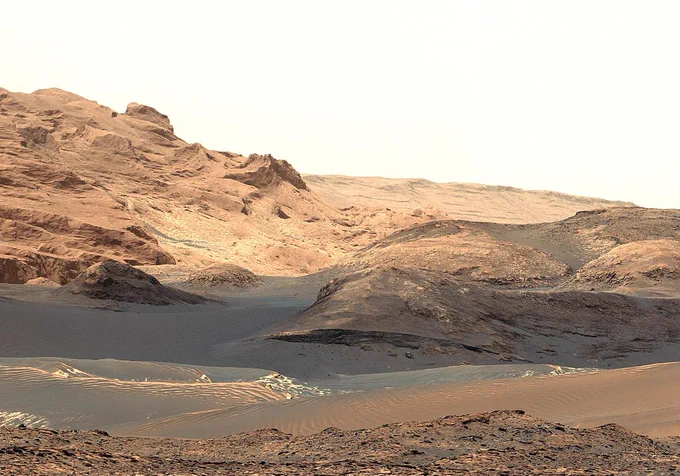 A desert landscape with dunes of sand and dust in the foreground, and craggy sandstone hills on the horizon.