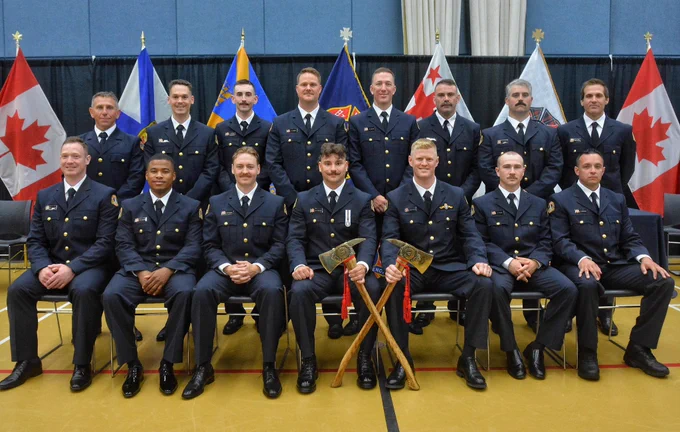 Group photo of the HRFE June 2025 Career Recruit Firefighter class at their graduation ceremony. Firefighters are in their dress uniform, with flags behind them and two in the front row are holding ceremonial axes.