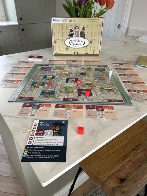 Legless in London, the board game, laid out ready to play on a worktop. The board depicts the streets of Victorian London with several locations on it. Around the board are locations cards. Also on the board are pawns and victory tokens. Behind the board is the game box with images of artificial legs and prosthesis users on it.