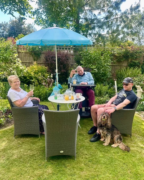 3 people and a dog sit around a table in a luscious green garden under a parasol. 
