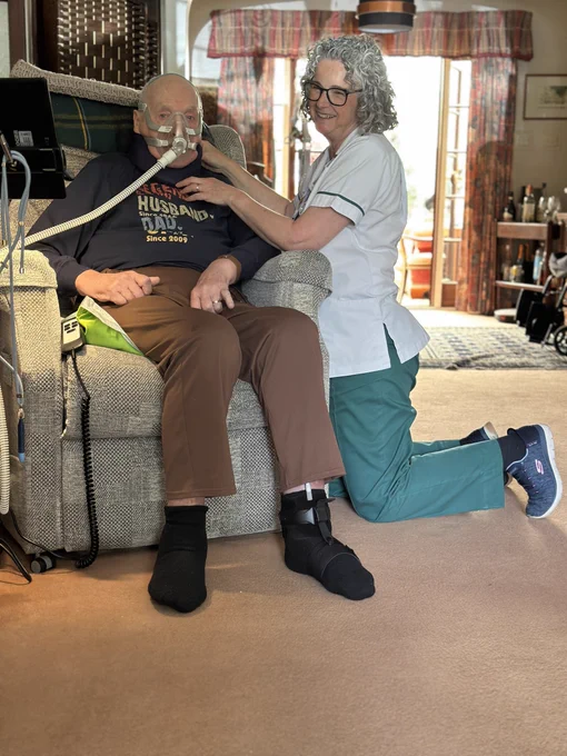 Heather kneels next to John, who is sat in a chair wearing an oxygen mask. 