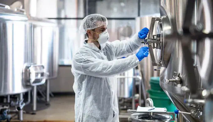 Technician in protective gear inspecting stainless steel equipment in a modern food processing facility, ensuring quality control of food and beverage ingredients.