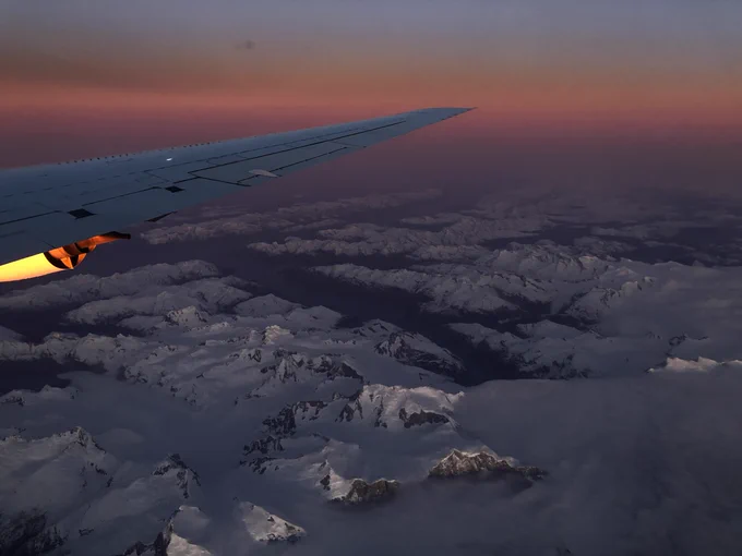 The wing of NASA's DC-8 plane, seen out the window against a vivid sunset. The plane is flying over rugged Patagonian mountains, covered in glacial ice. Dark water carves thick channels between the snowy mountains. The sunset casts a faint pinkish glow over the entire scene, which is dimly lit. In the distance, the sunset paints in the sky in three distinct bands, dark purple near the horizon, peachy pink in the center, and a dusky blue higher up.