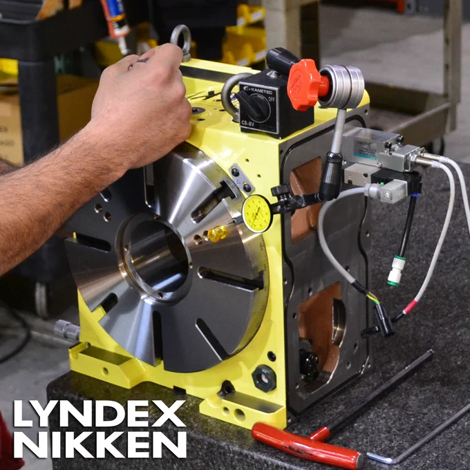 A close-up image of a person's hand adjusting a yellow and silver CNC rotary table, with various gauges and wires visible. The "LYNDEX NIKKEN" logo is visible in the bottom left corner.