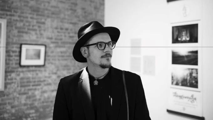 Black and white photo of Luke Townsend wearing a hat and glasses, standing in an art gallery with photographs displayed on a brick wall.