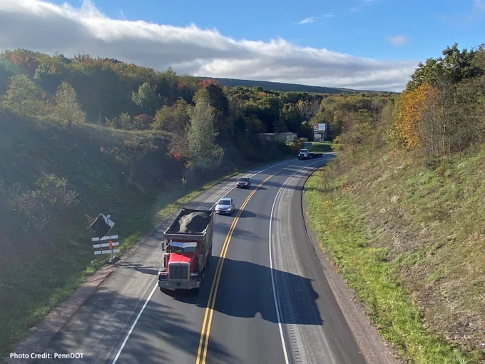 A scenic view of a highway winding through a hilly landscape with colorful autumn foliage, under a partly cloudy sky. Several vehicles, including a truck, are visible on the road. Photo Credit: PennDOT.

