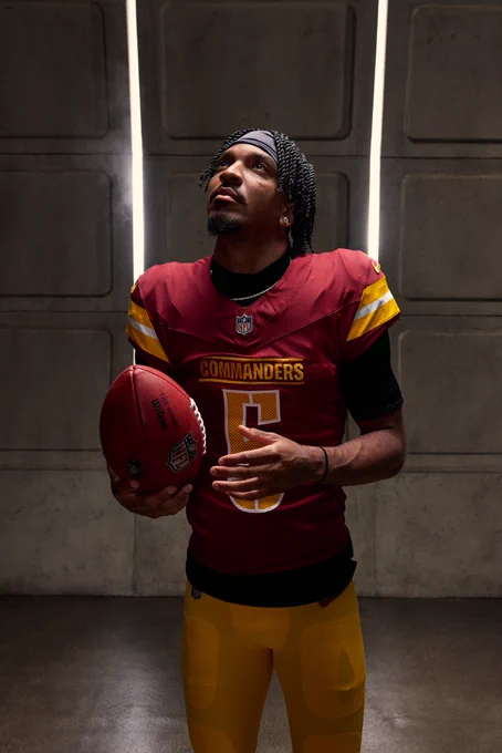 Photo of Jayden Daniels at media day. He is wearing a burgundy jersey and gold pants and is holding the football. The background looks like a Metro station.