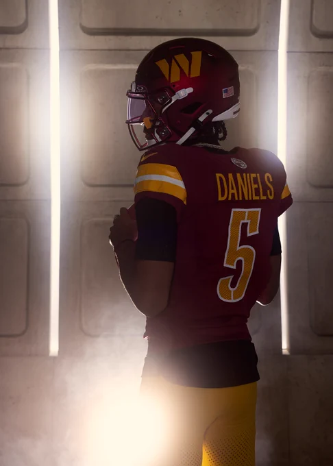 Photo of Jayden Daniels at media day. His back is to the camera and he is wearing a burgundy jersey and gold pants. The background looks like a Metro station.