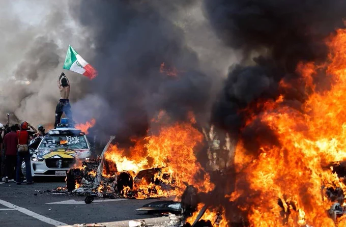 On top of a destroyed car, a man waves the flag of Mexico. Additional torched cars with heavy black smoke surround the man and others.