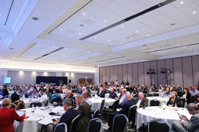 Conference audience sitting at round tables. There are people standing at the back of the room.