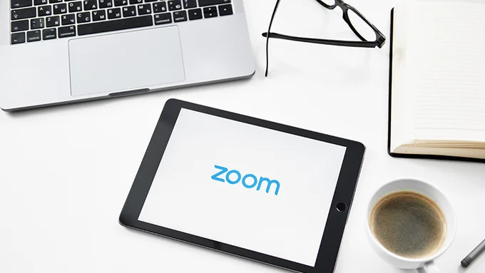 Photo shows tablet displaying the Zoom logo, cup of coffee, book, glasses and open laptop