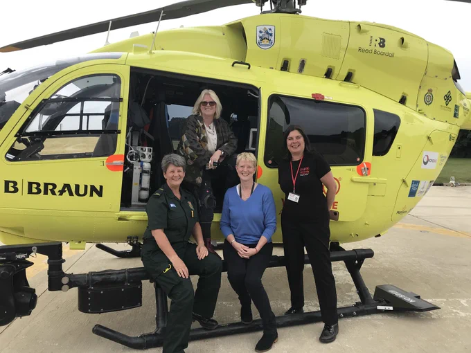 Three women stand together in front of a helicopter, smiling and dressed in casual outdoor attire.