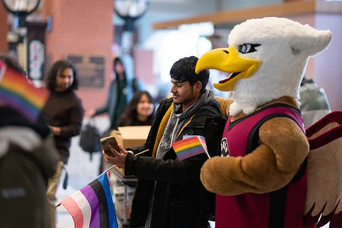 A Griffin mascot posing for a picture with someone as they hold small rainbow flags. 