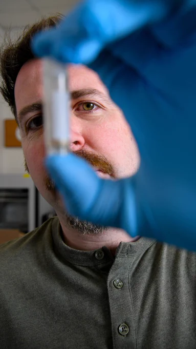 A photo of David Duffy wearing a green buttoned shirt and holding up an environmental sample wearing blue gloves on his hands towards the camera. The background includes some indistinct objects in a lab.