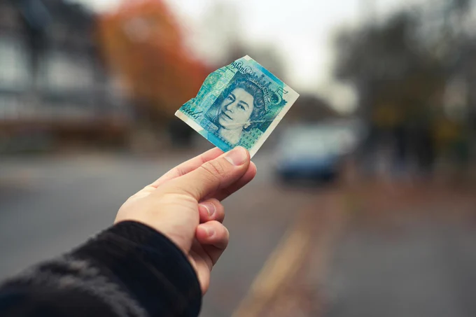 A photograph showing an outstretched handing holding a torn £5 note on a street with a blurred background. 
