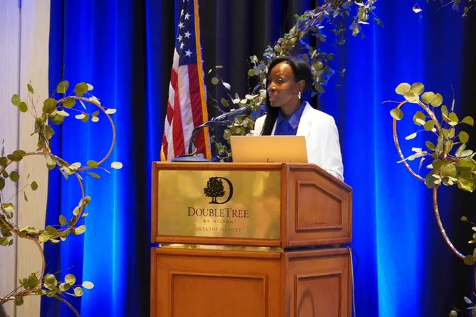 A person delivering a speech at a podium labeled DoubleTree by Hilton, with an American flag in the background and decorative plants around the stage.