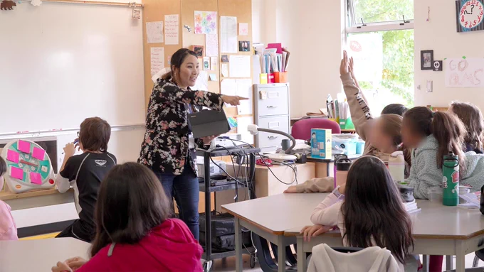 A woman holds an iPad at the front of the classroom. Students look towards her.  One child raises her hand. The woman points to her.