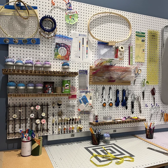 A well-organized craft station with various tools and supplies. Features include shelves holding jars of paint, a pegboard with hanging scissors and rulers, a desk with drawing materials, and containers of pens and brushes. A cutting mat and guides are laid out on the work surface.