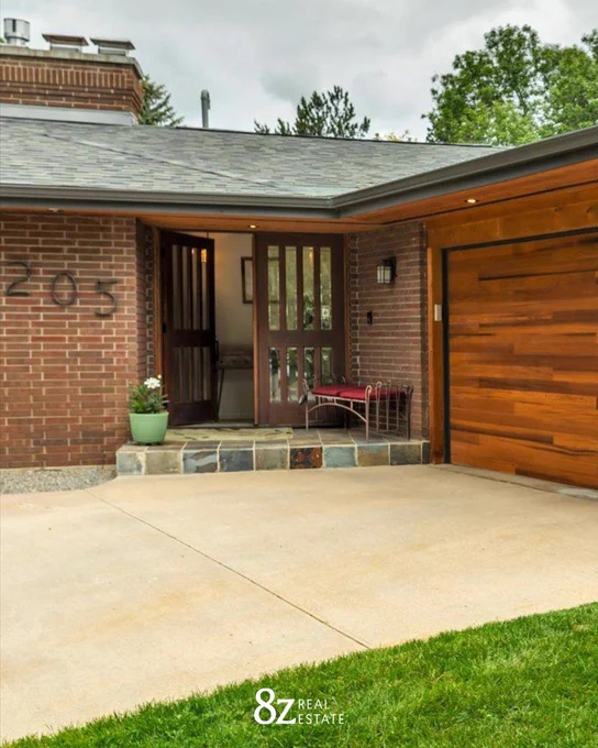 Entrance of a brick house with a wooden garage door adjacent to a covered porch area, featuring a small bench and decorative plants. The logo of 8z Real Estate is visible.