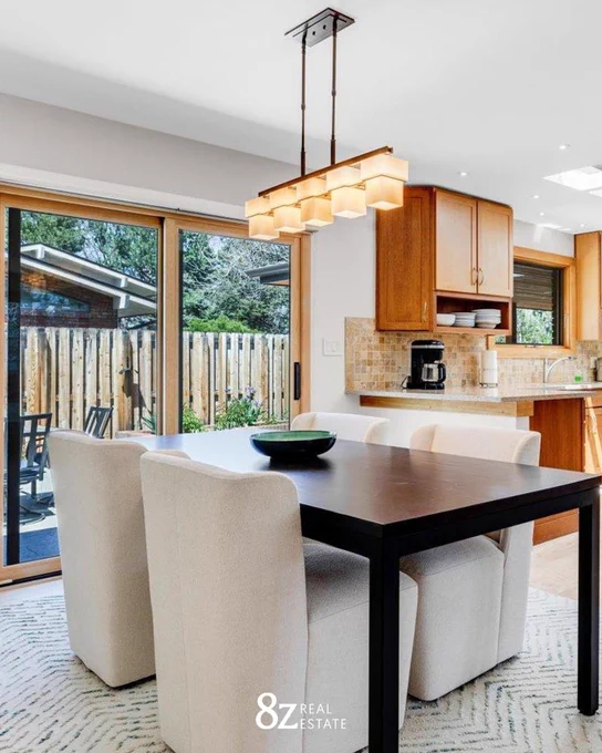 Modern kitchen interior featuring a dining table with chairs, wooden cabinets, and large windows overlooking a backyard. The scene includes a hanging light fixture and is branded with "8z Real Estate.