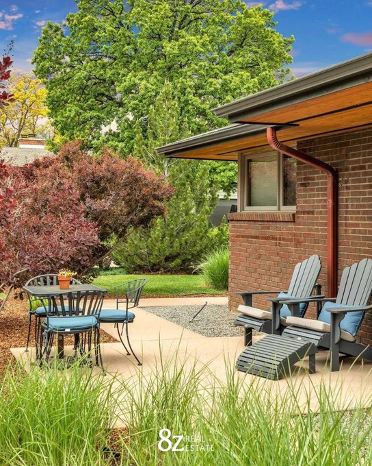 Cozy outdoor patio area with two Adirondack chairs and a small table, surrounded by lush greenery and trees, next to a red brick house. The image includes the logo of 8z Real Estate.
