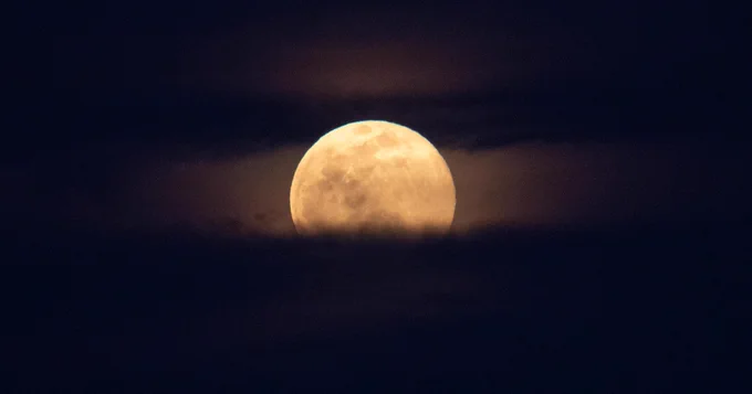 A large, golden Moon peeks out through banks of clouds and fog in the night sky. Craters and other features are visible on the glowing Moon. Credit: NASA/Joel Kowsky