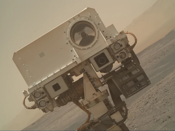 A portion of a selfie is shown with the Curiosity Mars Rover angled to the side of the image. The head is coming in from the right and tilted toward the left with its rectangular head taking up most of the image.