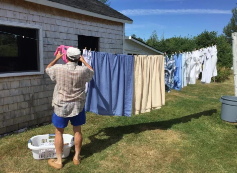 A person is hanging laundry on a clothesline outside a small house on a sunny day.