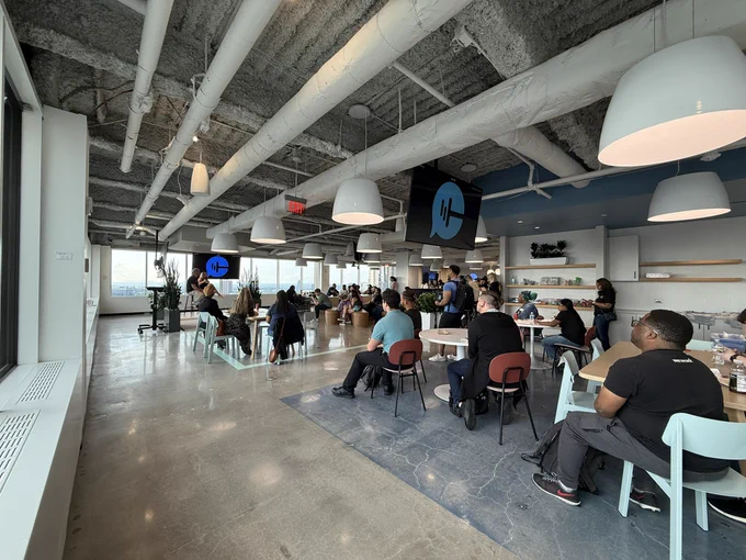 A modern, open-layout office space filled with people seated at various tables, attentively listening to a presentation. Large windows line the left side of the room with a scenic city view. Exposed ceilings with white ducts and hanging pendant lights create an industrial aesthetic.
