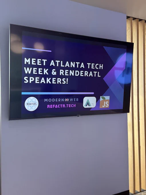 A digital screen mounted on a wall displaying the text “Meet Atlanta Tech Week & RenderATL Speakers!” with logos for ReactATL, Modern Web, Refactr.Tech, ATL, and JavaScript beneath the announcement. The background of the screen is a sleek mix of purples and blues with abstract shapes.