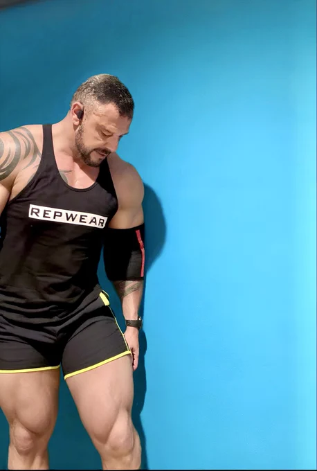 A muscular man wearing a black REPWEAR tank top and black shorts with yellow trim stands against a vibrant turquoise wall. He looks down, showcasing his defined physique, with visible tattoos and a black elbow sleeve on one arm.