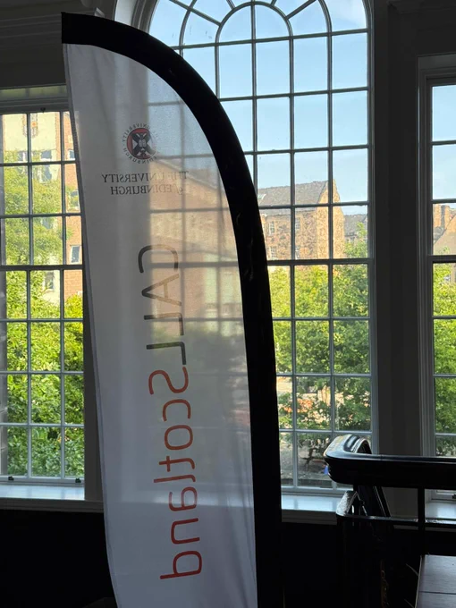 Photograph of the view from the tops of the stairs in Patersons Land at the University of Edinburgh. A banner with the text 'CALL Scotland University of Edinburgh' is in the foreground with  a tall arched window with paned glass in the background. The view through the window shows Moray House library surrounded by trees and bright blue sky in the sunshine.
