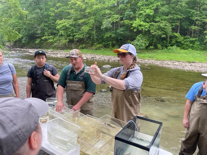Wetland Project Manager Carlyn Rocazella, wearing a yellow-rimmed hat, presents a live Silverjaw Minnow to a gathered group during a native fish identification activity.