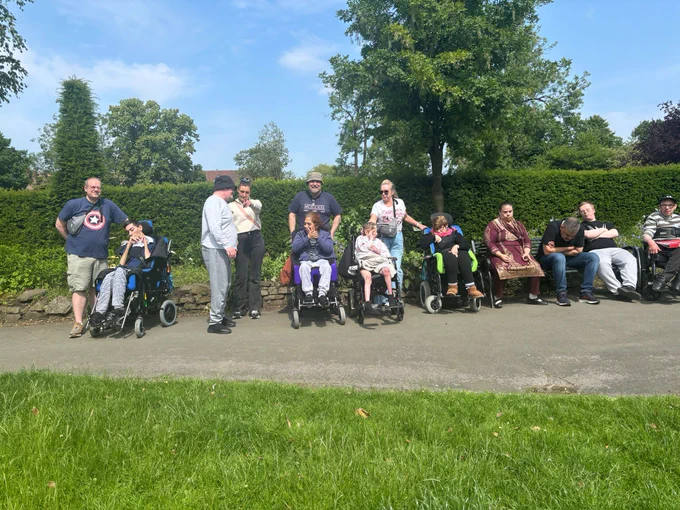 A group of people, some stood up and some wheelchair users, stood in a line on a gravel path in a park, with a grassy foreground and trees and blue sky in the background. 