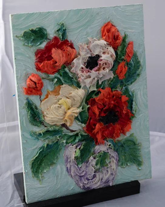 

A sculptural painting featuring vibrant 3D flowers, created with a palette knife. The artwork showcases red, white, and yellow flowers with textured green leaves, set against a light blue-green background, and is displayed on a black stand.