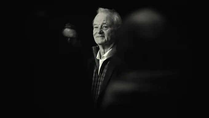 Soft-focus black-and-white portrait of Bill Murray, faint smile, light catching his face while background and bystanders fade into darkness.