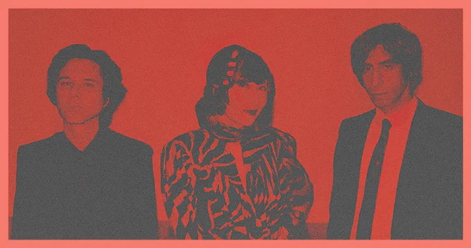 Three members of the band Yeah Yeah Yeahs, standing side by side against a red background.