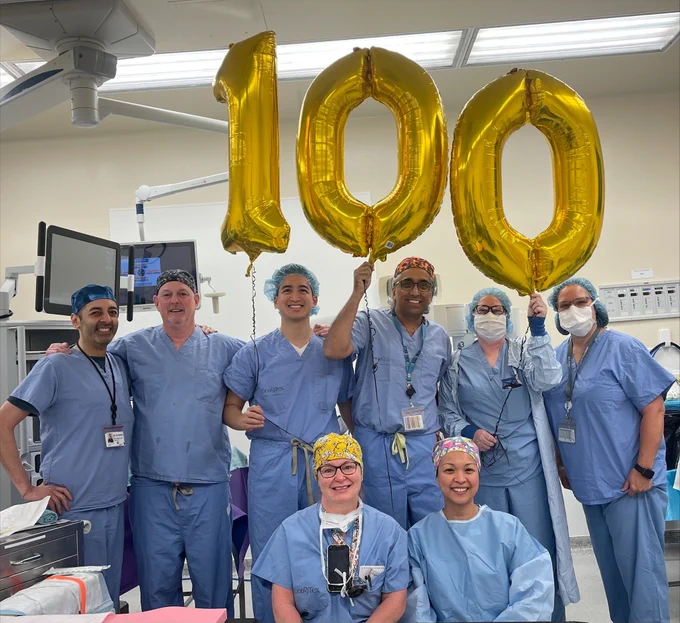 Eight members of the surgical team dressed in blue scrubs and standing in the surgical suite. They are holding up three gold foil balloons that read: "100."