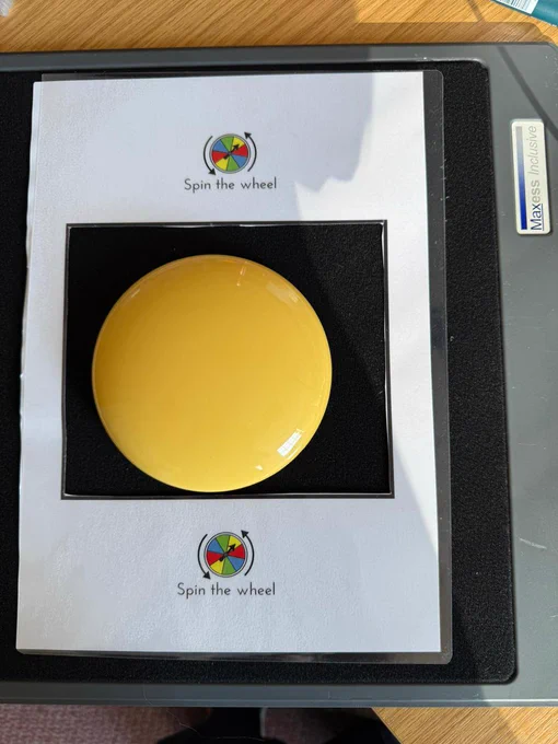 A photograph of a a large yellow wireless access switch on a black velcro tray. Above and below the switch is text that reads 'spin the wheel' with a picture symbol above it.