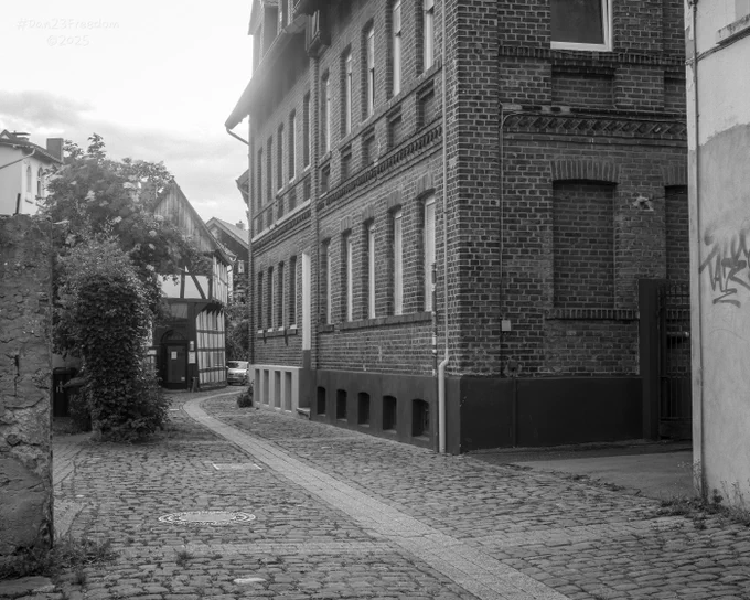 This monochromatic image transports the viewer to a narrow, winding street paved with cobblestones, flanked by an imposing brick building on the right and glimpses of older, timber-framed houses further down the lane on the left.