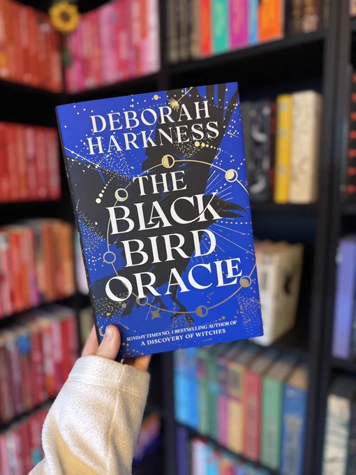 A person holds up a hardcover copy of The Black Bird Oracle by Deborah Harkness in front of a wooden bookcase filled with books. The book features a black cover adorned with gold foil designs, including a raven and celestial motifs.