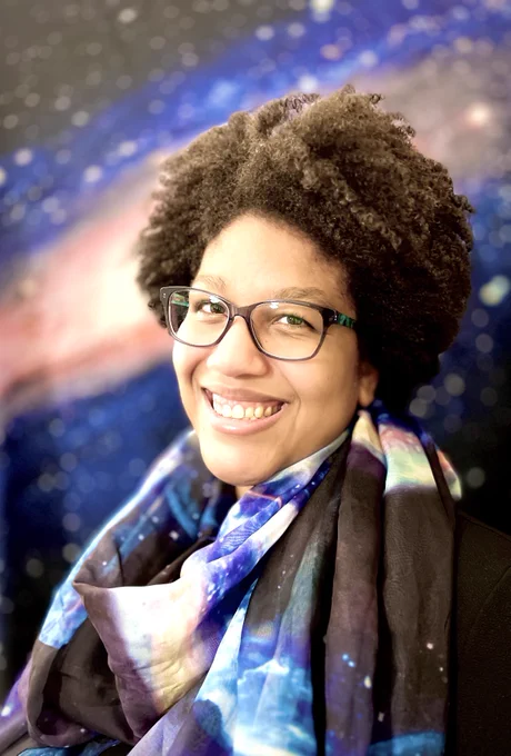 A smiling woman with short, curly hair wears glasses and a colorful galaxy-themed scarf. She is posed in front of a vibrant space-themed background that features stars and a nebula, giving the image a cosmic feel.