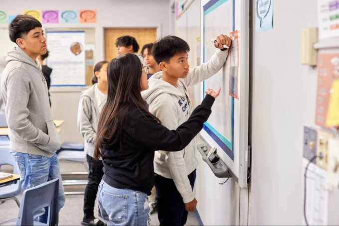 Students in a classroom facing and writing on a whiteboard on the wall.