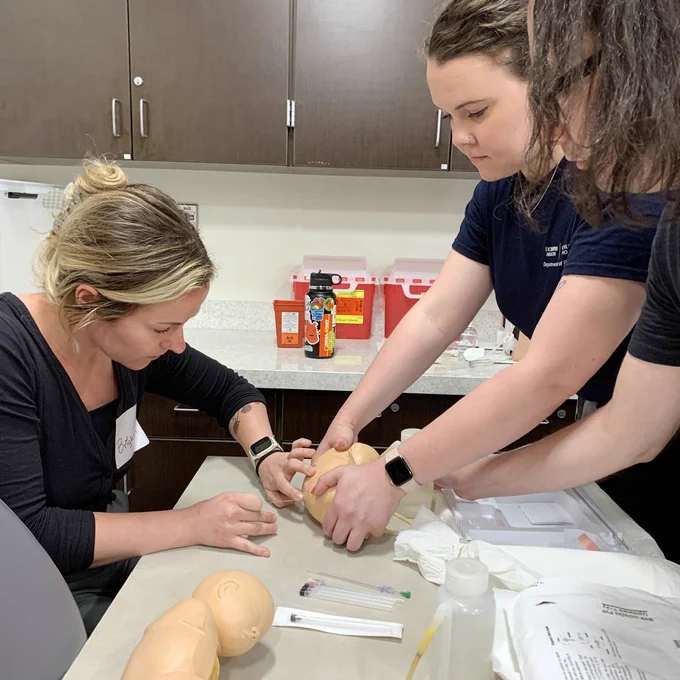 An intern practices a spinal tap on an infant simulation manikin as two instructors assist.