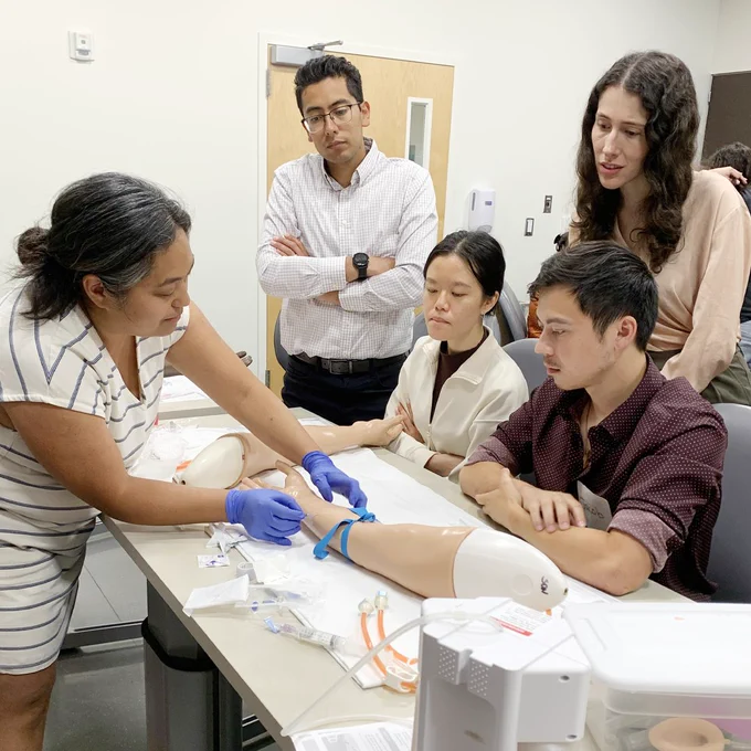 A faculty member instructs four interns on proper IV insertion technique using a simulated manikin arm.