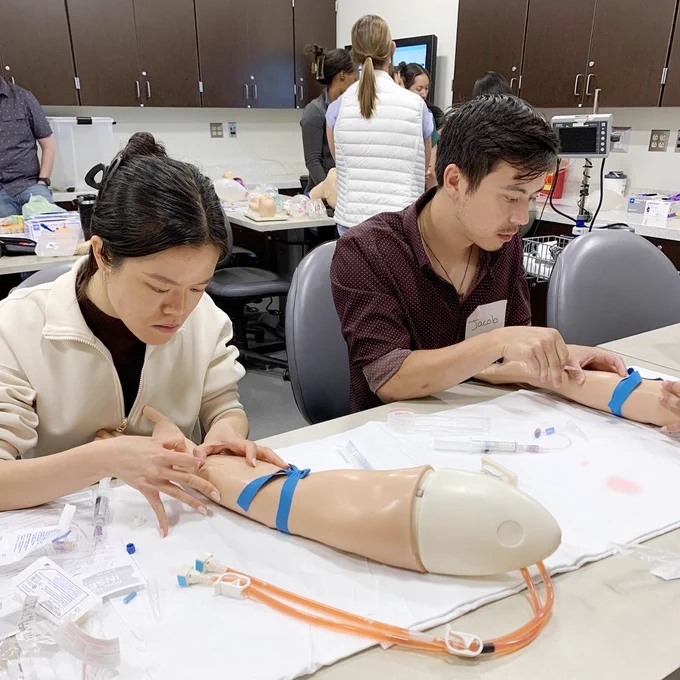 Two interns practice proper IV insertion technique using simulated manikin arms.
