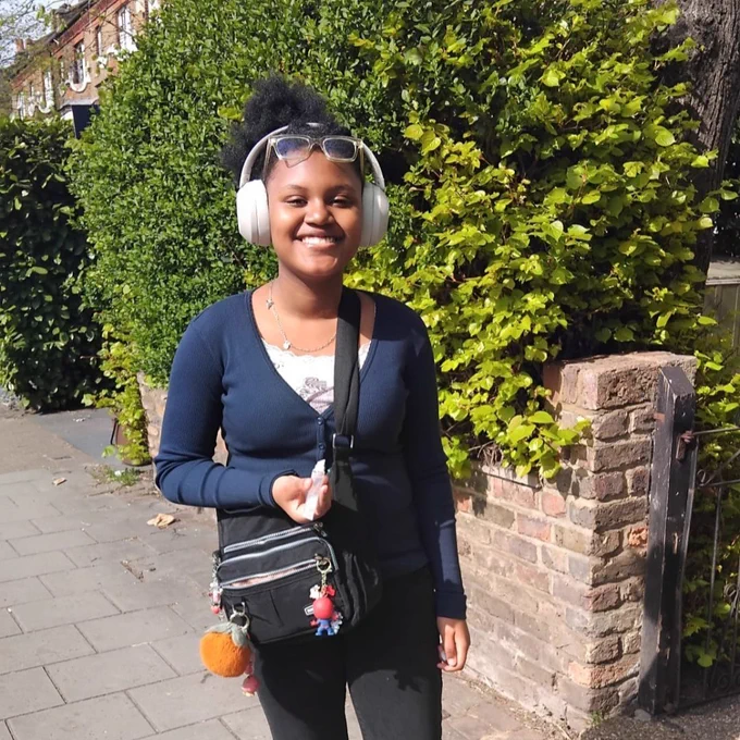 Sahara wearing headphones, and a navy cardigan over a white top, with a shoulder bag with some large keyrings on, smiling towards the camera whilst stood on a pavement